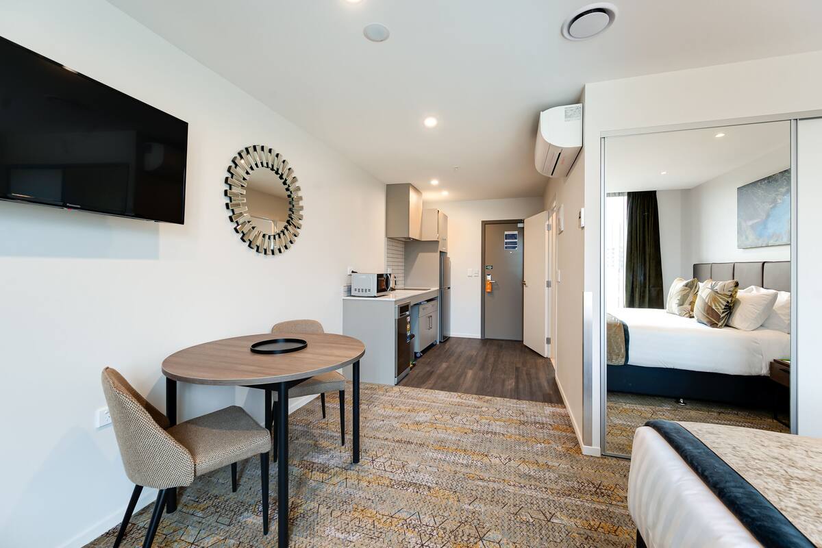 Ein modernes Hotelzimmer mit einem Bett, einer Essecke mit Tisch und Stühlen, einer Küche und einem an der Wand montierten Fernseher.