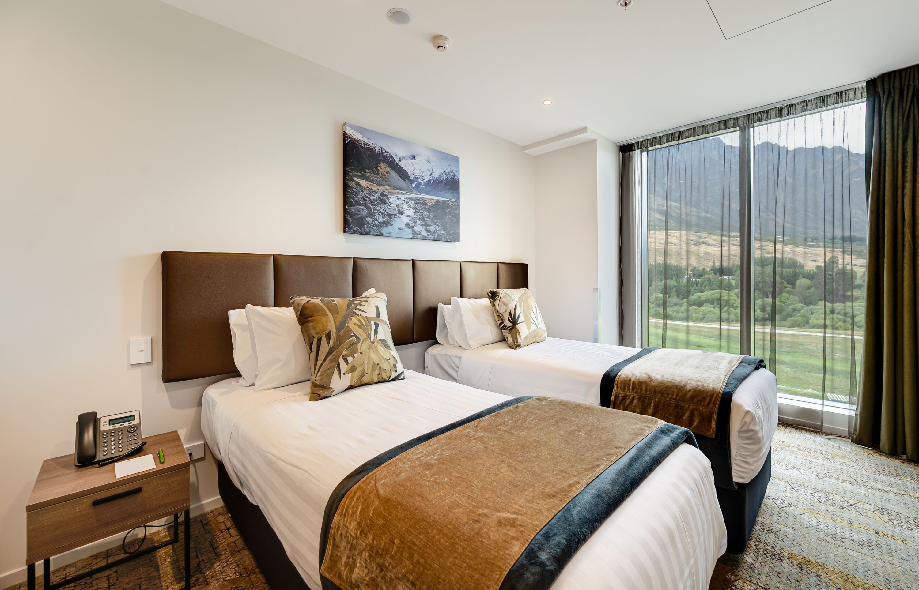 Une chambre d’hôtel avec deux lits, un tableau représentant un paysage de montagne, un téléphone et une fenêtre donnant sur un paysage verdoyant et des montagnes.