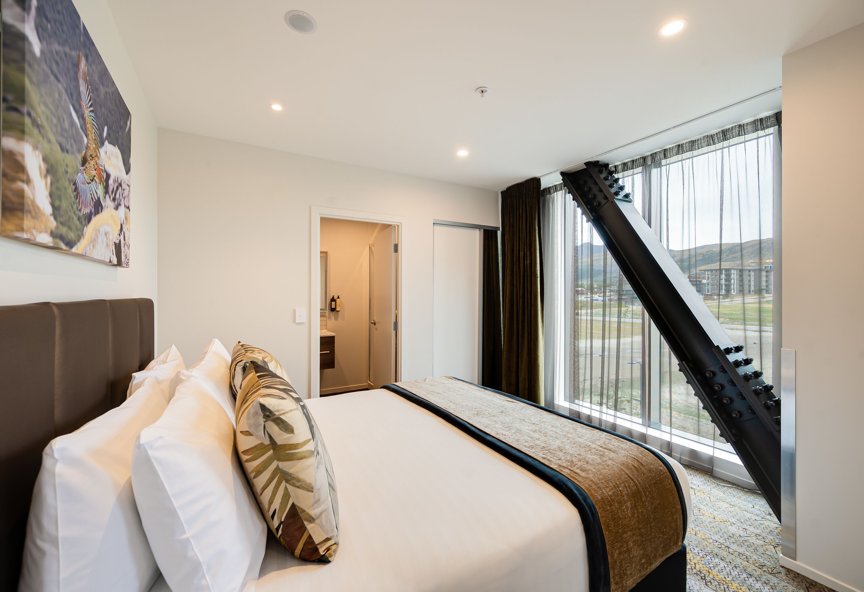 Une chambre d’hôtel avec un lit, une grande fenêtre avec des rideaux, une vue sur un paysage montagneux et une porte donnant sur une salle de bain.