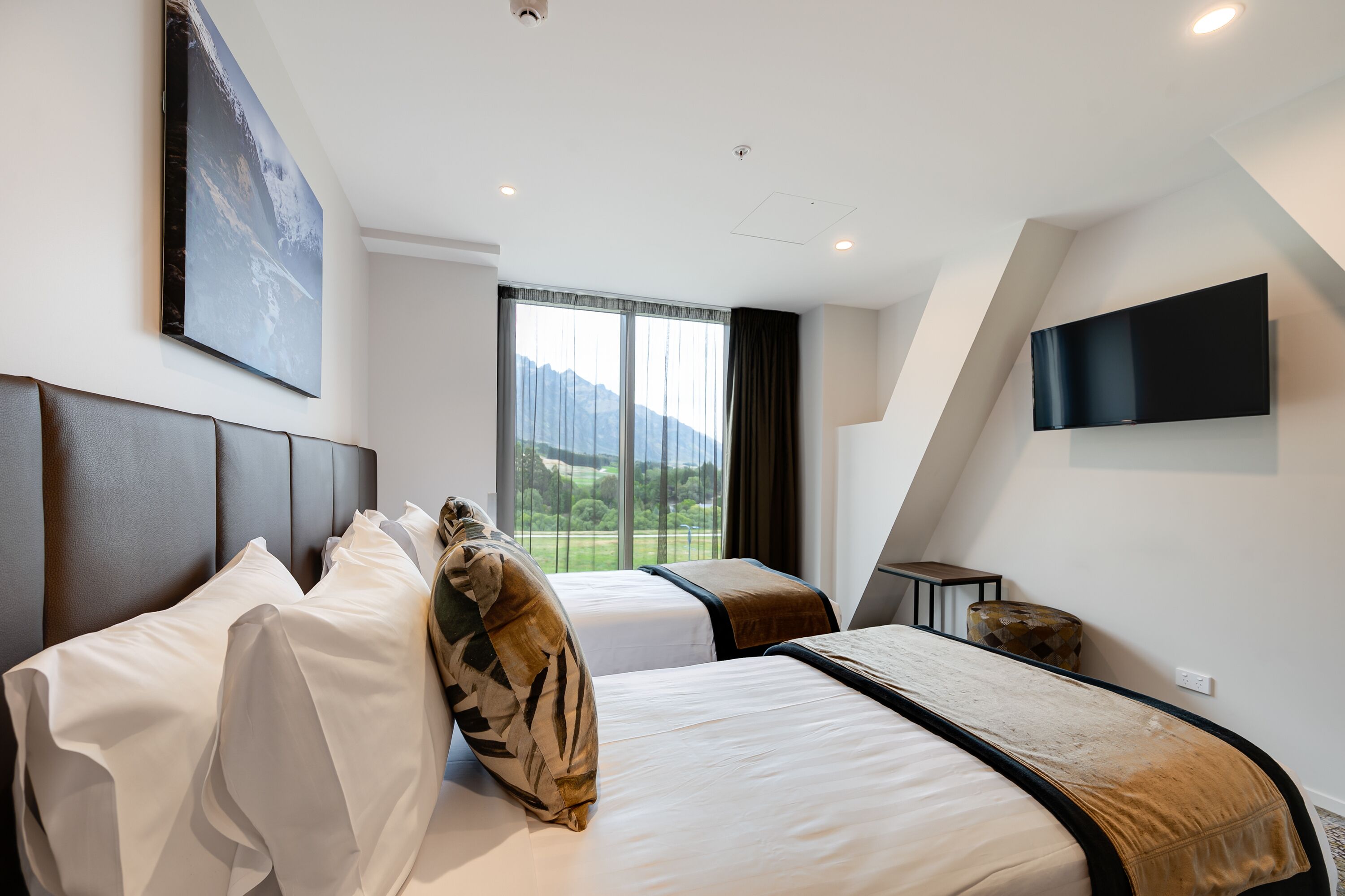 A modern hotel room with two beds, a large window with a mountain view, a flat-screen TV mounted on the wall, and a small side table.