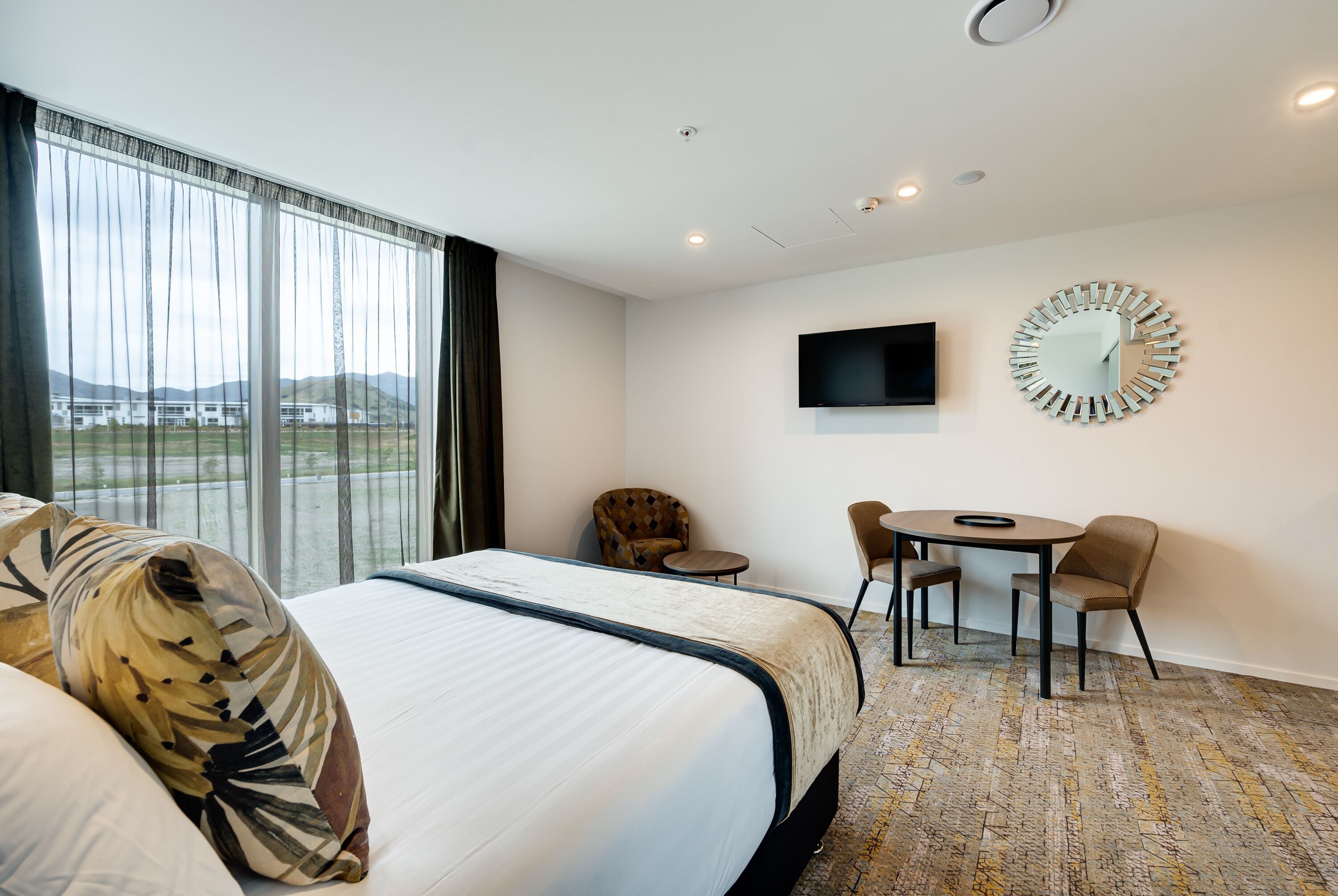 Een hotelkamer met een groot bed, een bureau met twee stoelen, een televisie en een decoratieve spiegel.