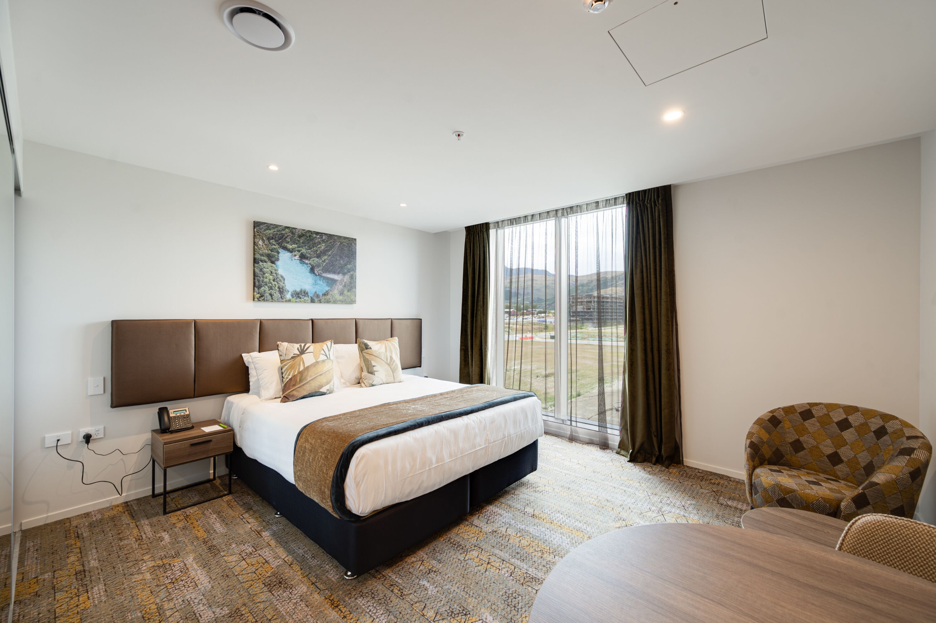 A hotel room with a large bed, a view of the outdoors, a dining table, and a patterned armchair.