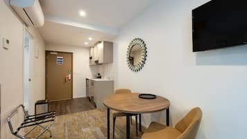 Una habitación moderna con una mesa redonda de madera, dos sillas de mimbre, un televisor montado en la pared y una zona de cocina al fondo.