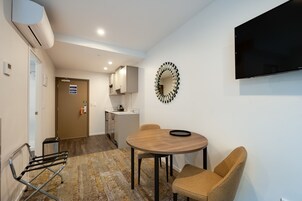 Una habitación moderna con una mesa redonda de madera, dos sillas de mimbre, un televisor montado en la pared y una zona de cocina al fondo.