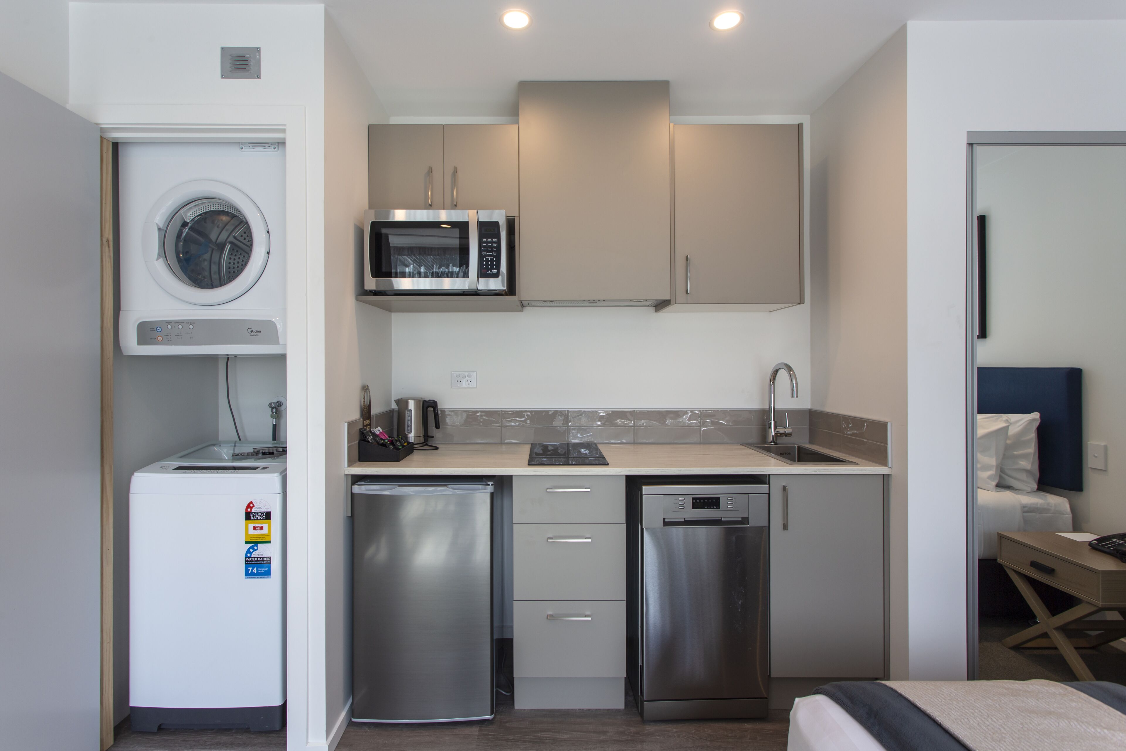 A compact hotel room with a kitchenette including a washing machine, microwave, refrigerator, and dishwasher.