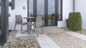 Un espace extérieur aménagé avec deux fauteuils en osier et une petite table, une porte vitrée donnant sur un jardin, et un arbuste taillé avec soin.