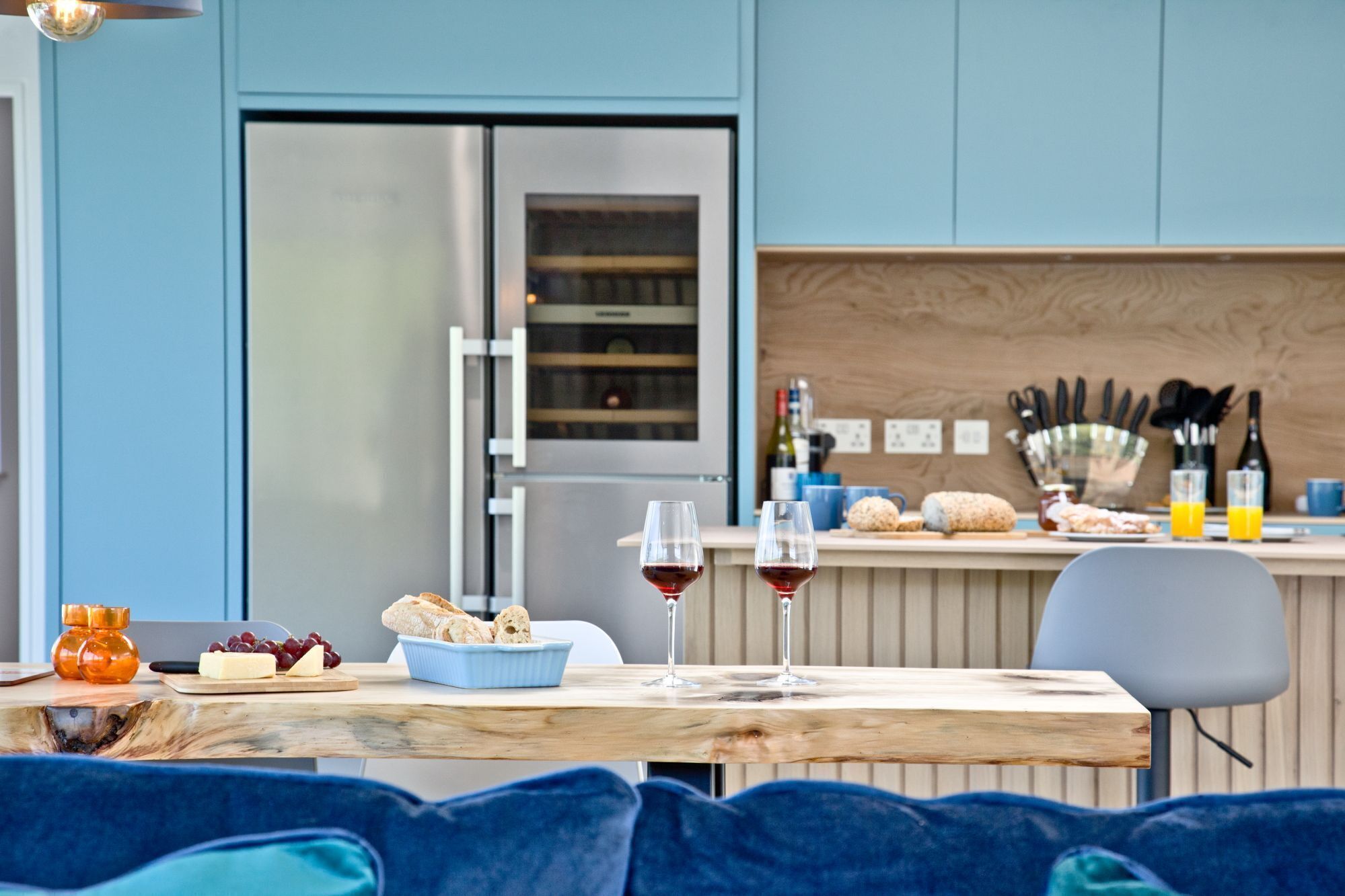 A modern kitchen with a wooden table set for a meal, featuring bread, cheese, grapes, and wine glasses.