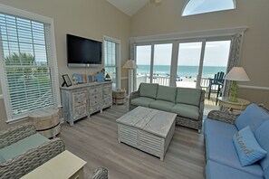 Amplia sala de estar con un gran ventanal con vistas a la playa, un televisor de pantalla plana sobre un mueble de madera, y una zona de estar cómoda con un sofá azul y un sofá verde.