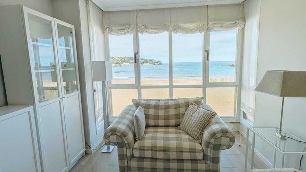 A room with a checkered sofa facing a large bay window overlooking a beach. - Beach apartment with stunning views (Noja)