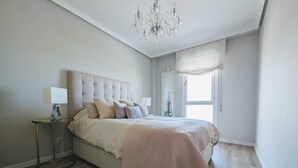 A bedroom with a large bed, a chandelier, a bedside table with a lamp, a window with sheer curtains, and a patterned rug. - Beach apartment with stunning views (Noja)