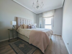 A bedroom with a large bed, a chandelier, a bedside table with a lamp, a window with sheer curtains, and a patterned rug.
