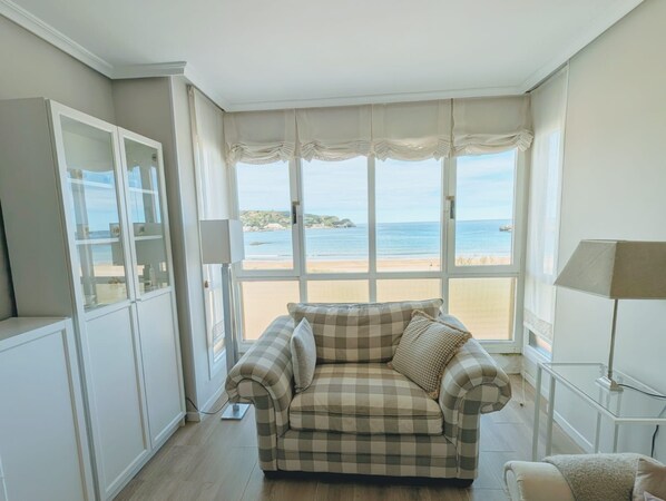 A room with a checkered sofa facing a large bay window overlooking a beach.