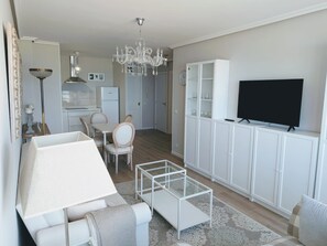 A modern living room with a sofa, a glass coffee table, a TV on a cabinet, a chandelier, and a kitchen in the background.