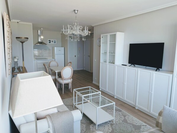 A modern living room with a sofa, a glass coffee table, a TV on a cabinet, a chandelier, and a kitchen in the background.