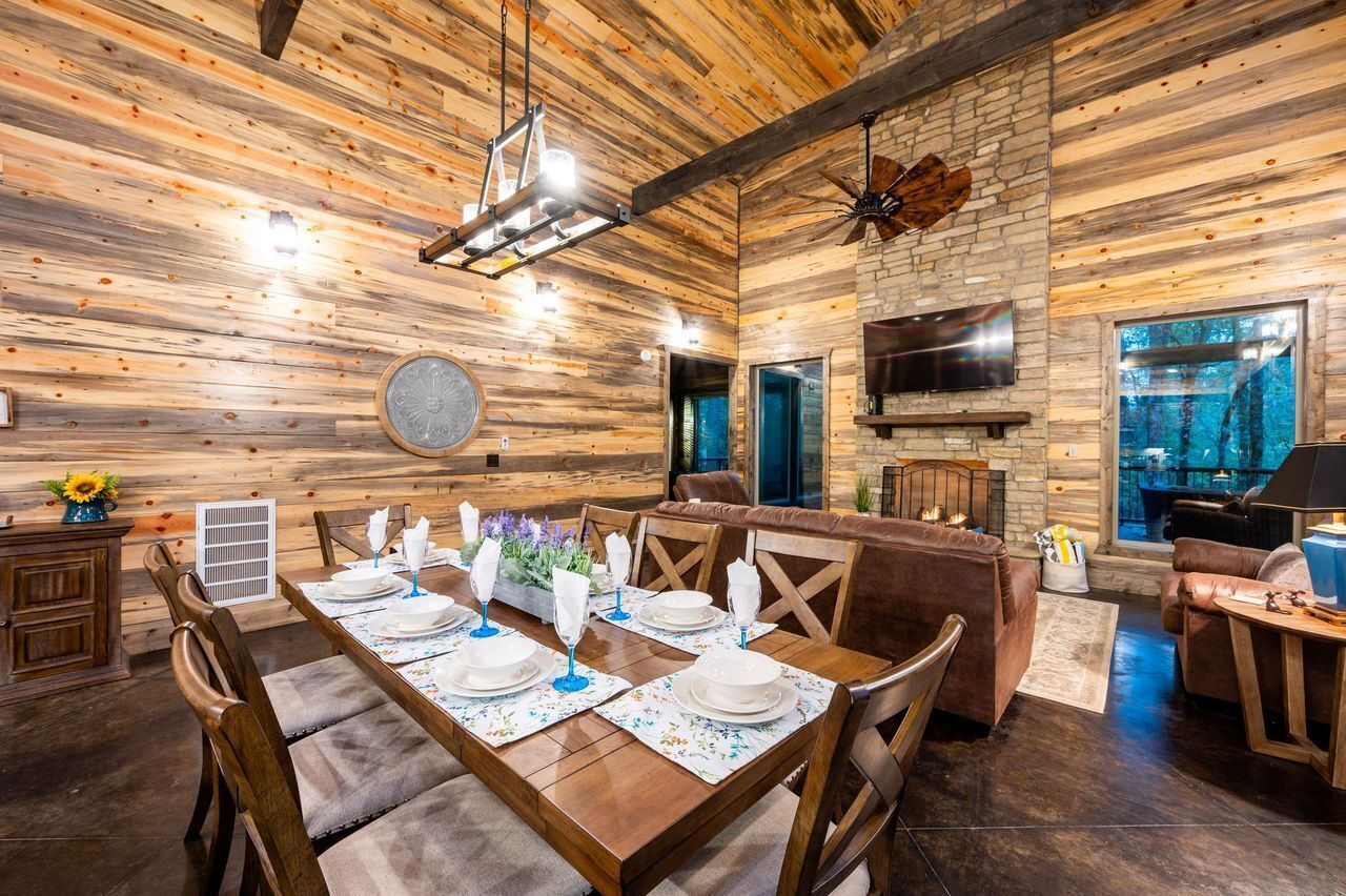 A rustic dining area with a wooden table set for six, a stone fireplace, and a large window overlooking a forest.