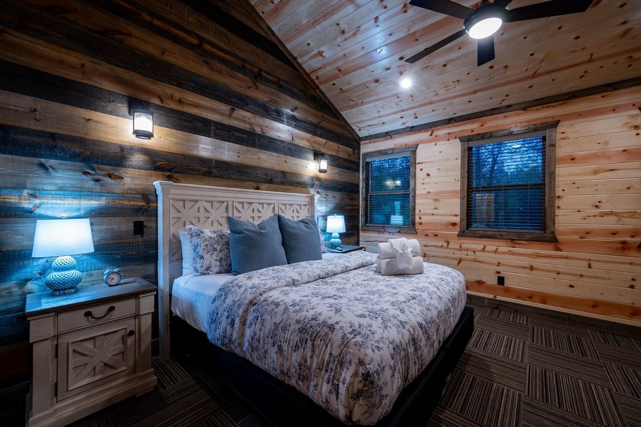 Un dormitorio de cabaña de madera con cama, mesitas de noche y un ventilador de techo.