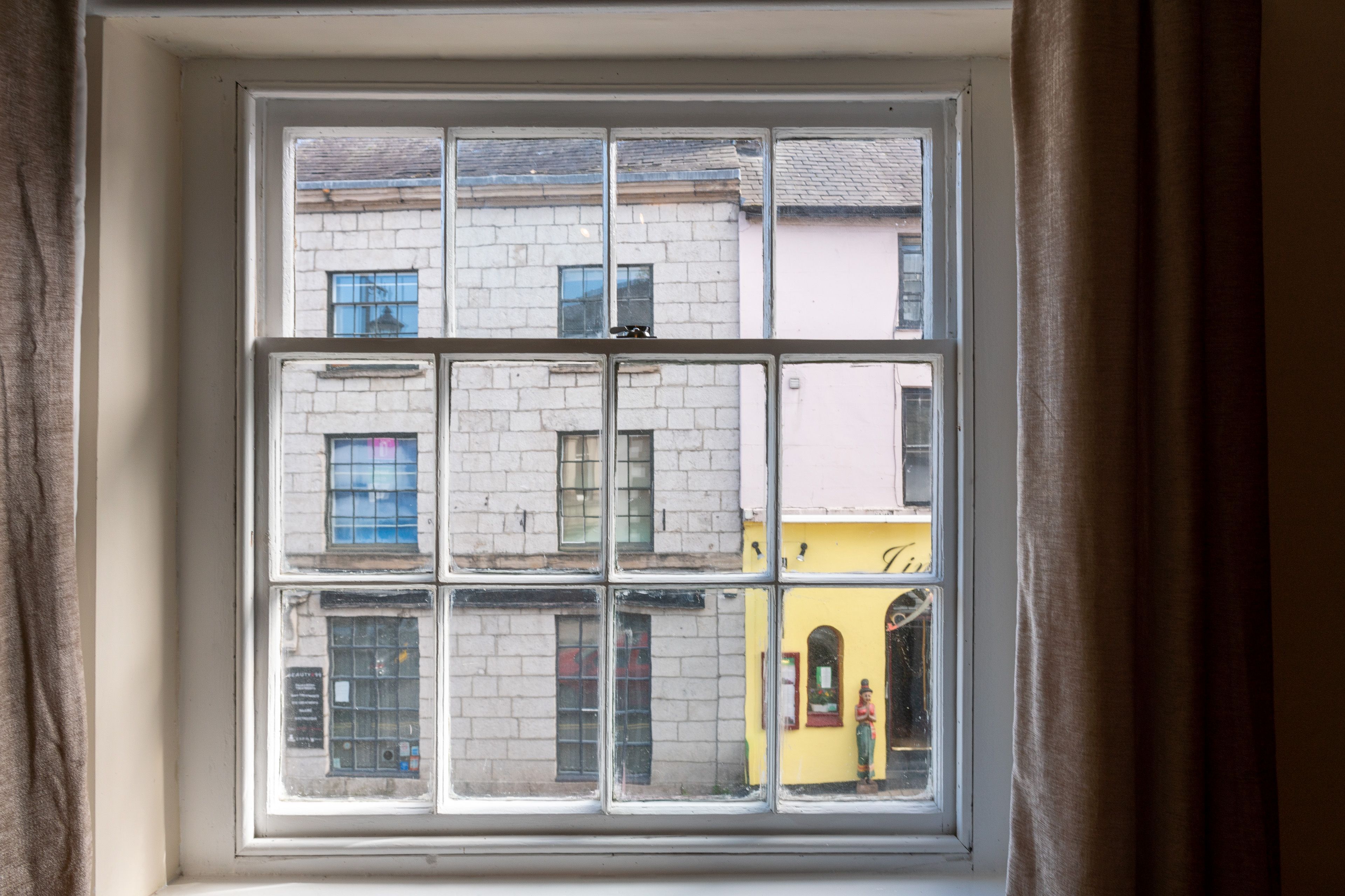 A window with a grid pattern, offering views of buildings with brick facades and a yellow building with a sign.