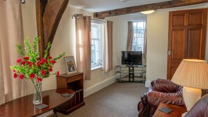 A living room with a wooden ceiling, a TV, a leather sofa, a lamp, a vase with flowers, and a window with curtains. - Highgate View Apartment -   Kendal Town Centre (Kendal)