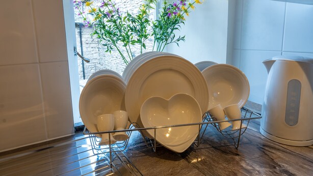 A kitchen with white plates and bowls on a rack, a white kettle, and a window with flowers. - Highgate View Apartment -   Kendal Town Centre (Kendal)