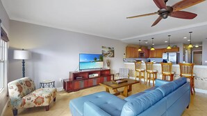 A living room with a blue sofa, a TV, a ceiling fan, and a kitchen area with wooden cabinets and yellow pendant lights. - Top Floor Waterfront Resort. Steps to the Beach,Pool, Hot Tub, Dock, Great View +Tickets to Fun! (Treasure Island)