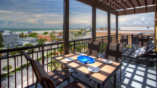 A balcony with a table set for dining, overlooking a beach and cityscape. - Top Floor Waterfront Resort. Steps to the Beach,Pool, Hot Tub, Dock, Great View +Tickets to Fun! (Treasure Island)