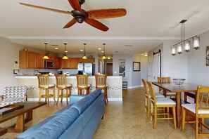 A spacious living room with a blue sofa, wooden dining table, and chairs, a kitchen area with wooden cabinets and a refrigerator, and a ceiling fan.