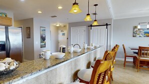 A kitchen with granite countertops, a refrigerator, and a dining area with wooden chairs and a table. - Top Floor Waterfront Resort. Steps to the Beach,Pool, Hot Tub, Dock, Great View +Tickets to Fun! (Treasure Island)