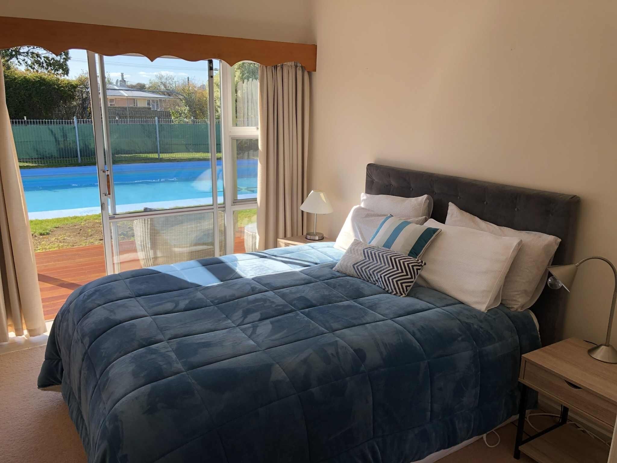Une chambre avec un grand lit, une table de chevet, une lampe et une vue sur une piscine, visible à travers une porte vitrée.
