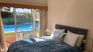A bedroom with a large bed, a nightstand, a lamp, and a view of a swimming pool through a glass door.