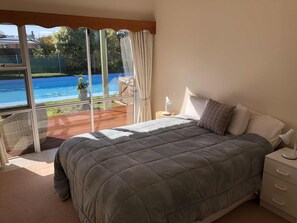 A bedroom with a large bed, a nightstand, a lamp, and a view of a swimming pool through a glass door.