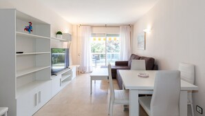 A modern, well-lit living room with a white sofa, a dining table, and a TV on a stand. There is a balcony with a view of greenery. - AirCon Apartment with Pool on the rooftop · UHC CENTRAL PARK 327 (Salou)