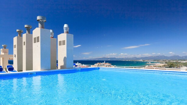 A swimming pool with clear blue water, lounge chairs, and a view of the sea and mountains in the background. - AirCon Apartment with Pool on the rooftop · UHC CENTRAL PARK 327 (Salou)