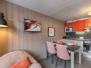 A modern living room with a sofa, a dining table with chairs, a TV, and a kitchen area with orange cabinets and a microwave.