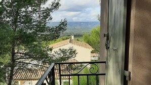 Un balcon doté d’une balustrade en métal, offrant une vue panoramique sur un paysage verdoyant, avec des arbres et des bâtiments situés au loin.