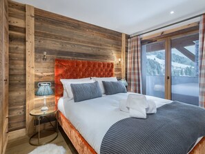 Quarto com cabeceira de madeira, cama grande com roupa de cama branca, duas almofadas, mesa de cabeceira com abajur, e vista para uma paisagem nevada através de uma porta de vidro.
