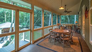 Eine überdachte Veranda mit Holztisch und -stühlen, großen Fenstern und einem Deckenventilator.