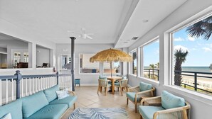 A spacious indoor area with a dining table and chairs, a sofa, and a large window offering a view of the beach.