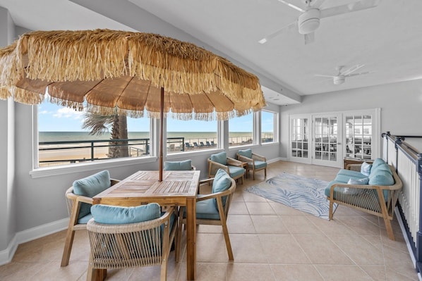 A spacious room with a large window overlooking the beach, a wooden table with chairs, and a thatched umbrella.
