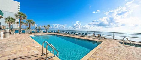 A pool area with a clear blue pool, lounge chairs, and a view of the ocean.