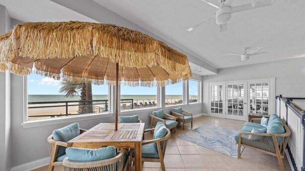 A spacious room with a large window overlooking the beach, a wooden table with chairs, and a thatched umbrella.
