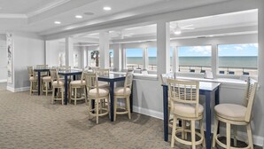 A spacious room with multiple tables and chairs, large windows with ocean views, and a ceiling with recessed lighting.