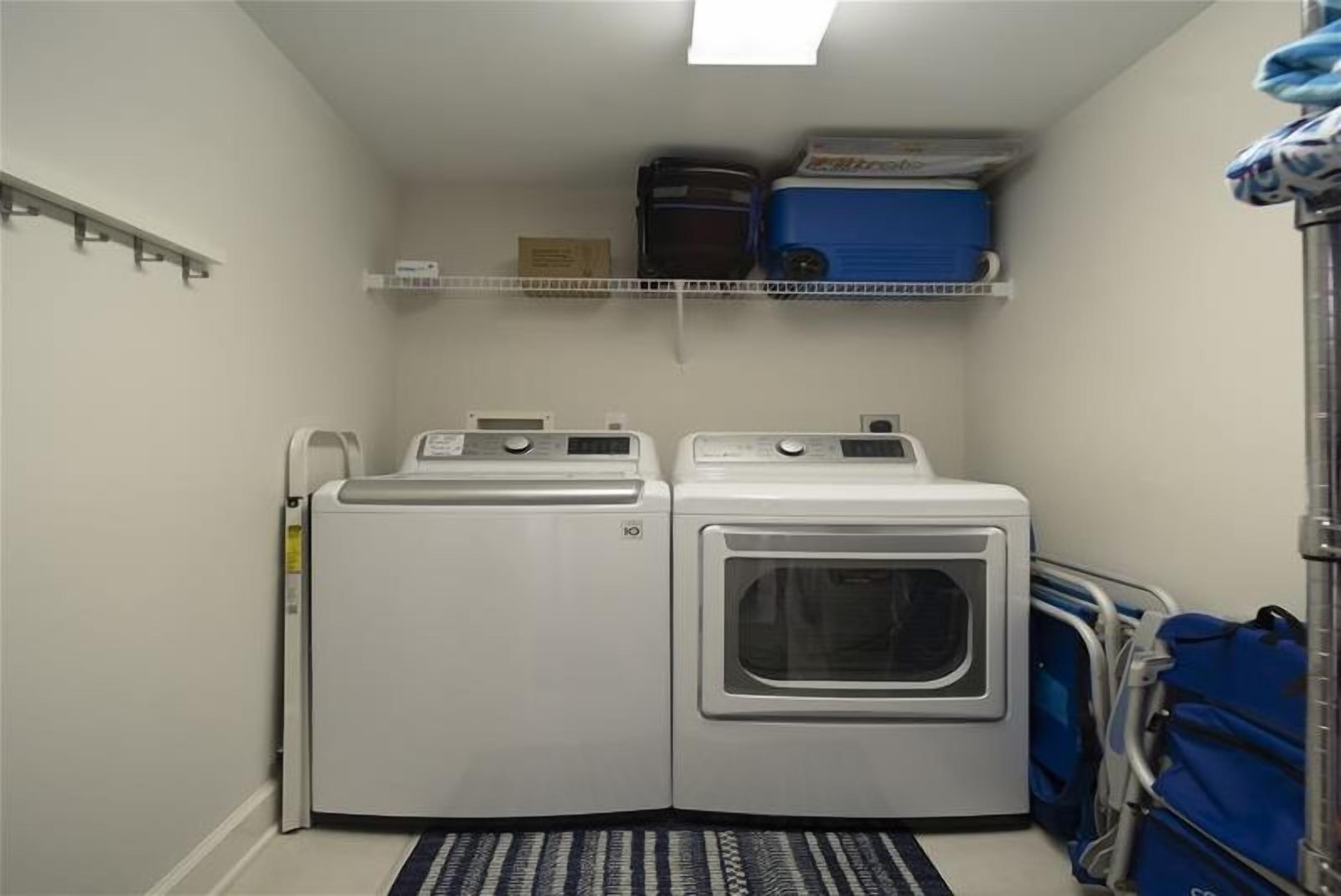 A laundry room with two stacked washers and dryers, a shelf above with folded laundry, and a blue luggage cart to the right.