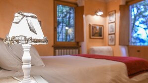 Un lit recouvert d’une couverture blanche et d’un plaid rouge, une lampe blanche ornée d’un ruban, et une fenêtre donnant sur des arbres.