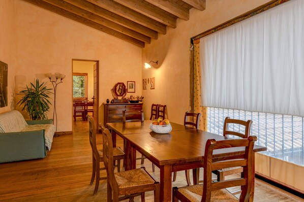 A dining area with a wooden table and chairs, a bowl of fruit, and a view through a window with curtains.