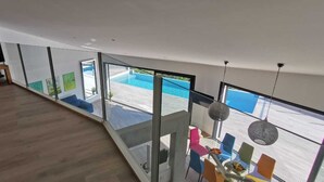 Uma sala de estar moderna com uma televisão grande, um móvel de entretenimento de madeira e um guarda-corpo de vidro com vista para uma piscina.