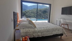 Quarto com uma janela grande que oferece vista para uma paisagem montanhosa e uma praia.