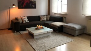 Un salon moderne avec un canapé foncé, une table basse blanche ornée de bougies, un pouf gris et un tableau accroché au mur.