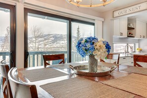 Un coin repas avec une grande fenêtre, un vase de fleurs et une cuisine en arrière-plan.