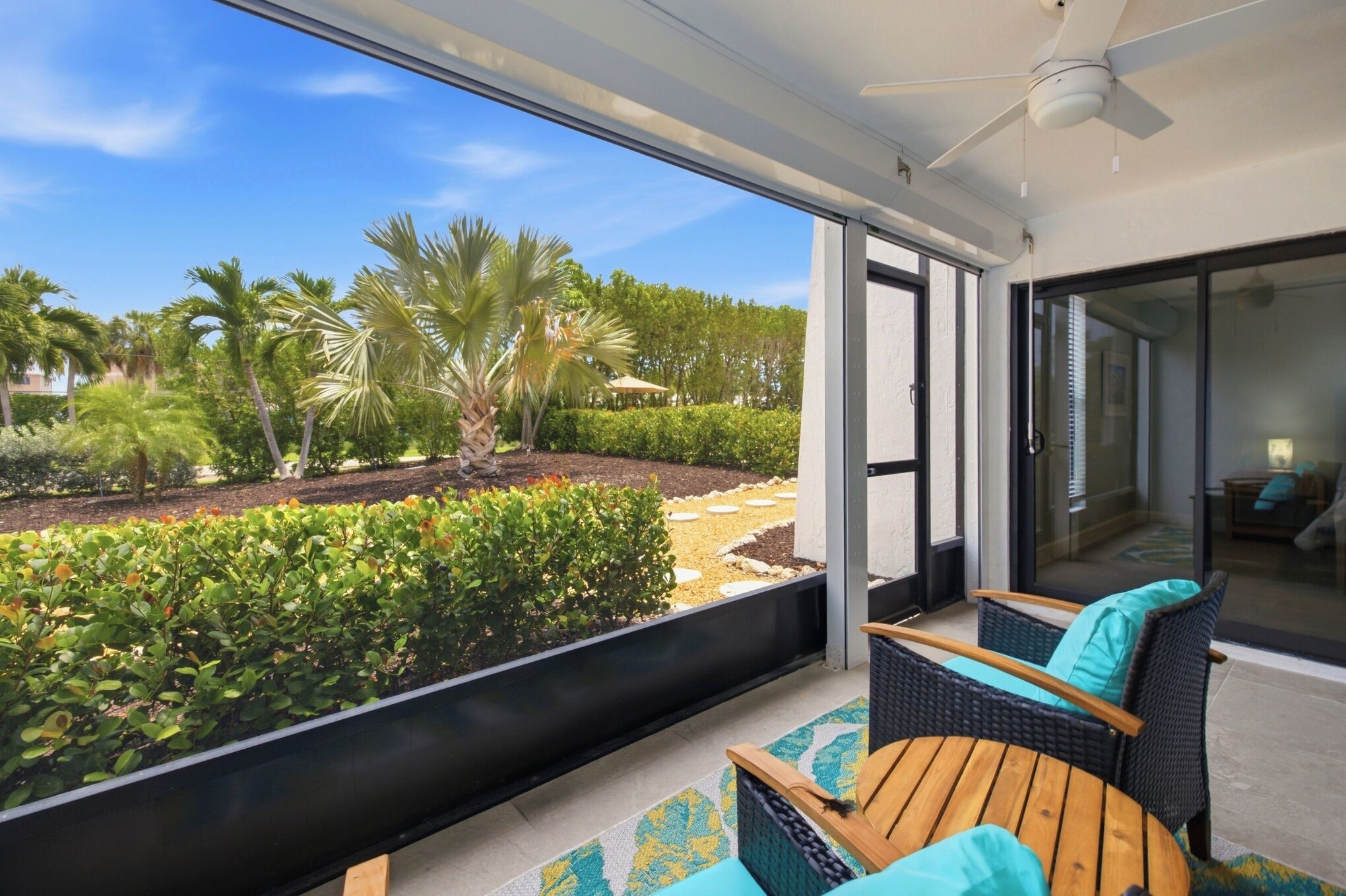 A balcony with wicker furniture, a ceiling fan, and a view of a landscaped garden with palm trees.