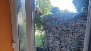 A view through a glass door into a courtyard with a stone wall, tiled floor, and some plants. - Vacation home Côtes d'Armor in the commune of Quessoy (Quessoy)